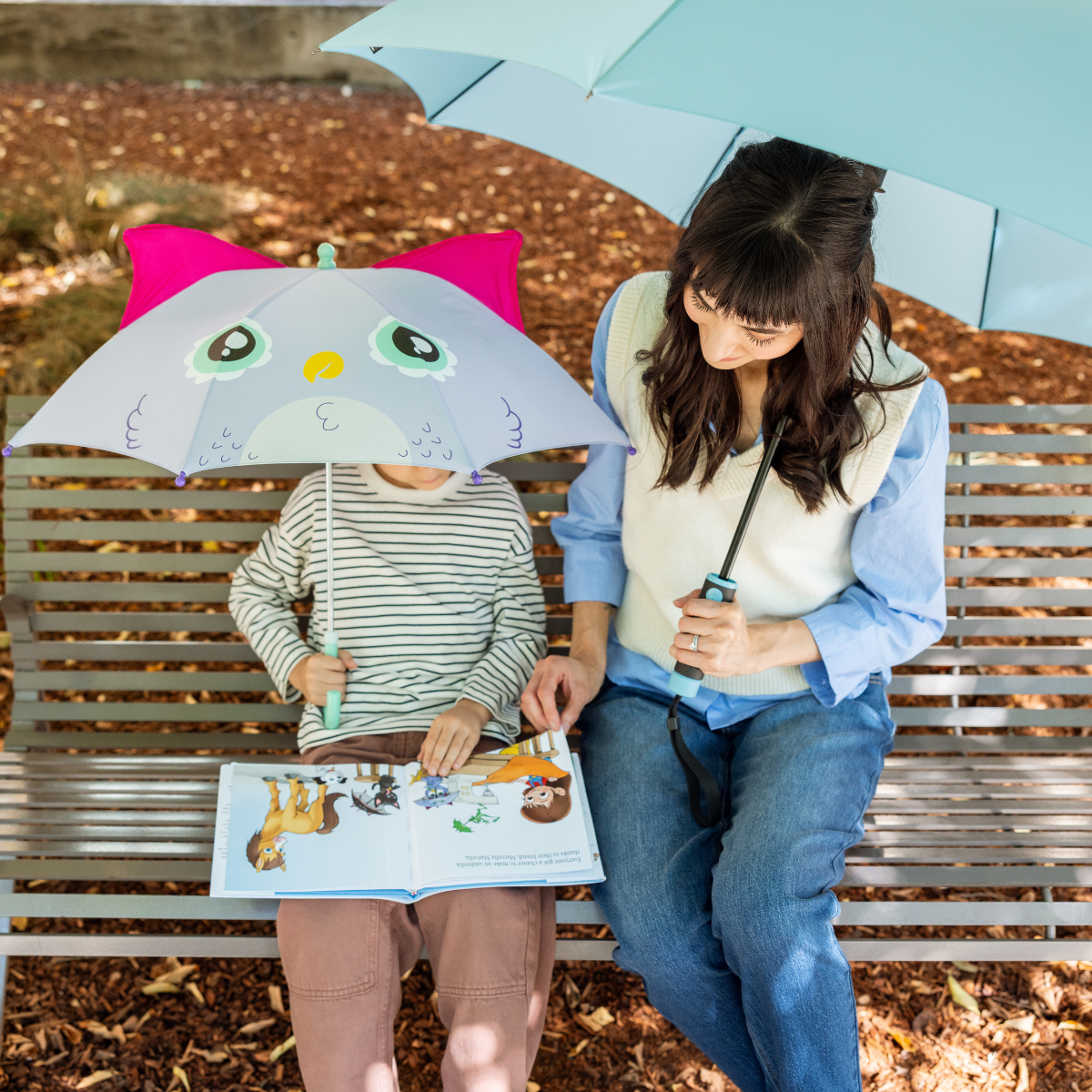 Two people sitting on a bench under umbrellas, one holding an open book.
