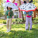 Two children holding animal-themed umbrellas in a grassy area with a house in the background.