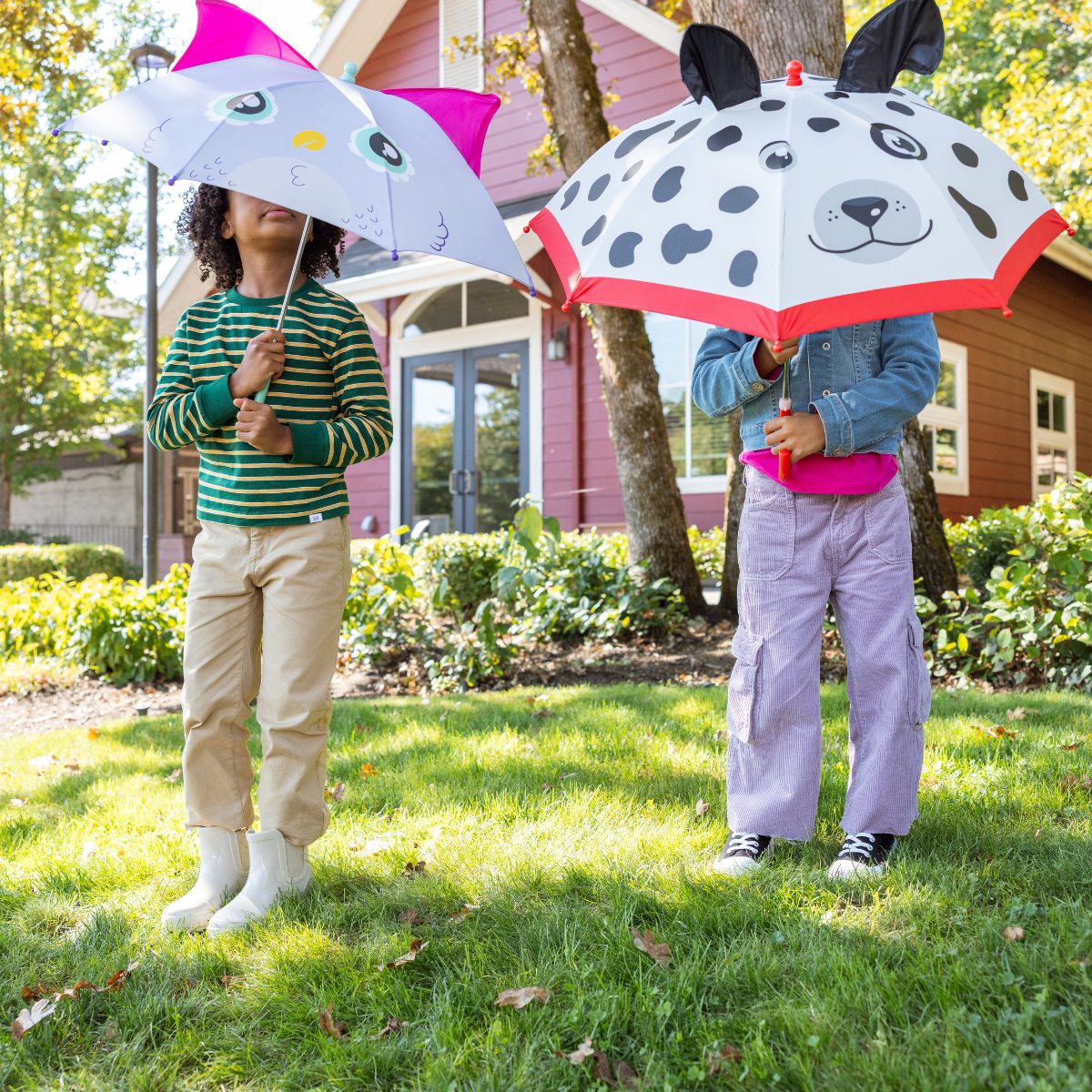 Two children holding animal-themed umbrellas in a grassy area with a house in the background.